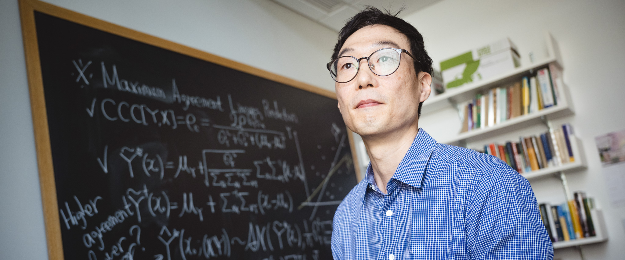Taeho Kim wearing a blue button-up shirt standing in front of a chalkboard with math problems on it.