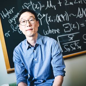 Taeho Kim wearing a blue button-up shirt and glasses sitting in front of a blackboard with math problems on it.