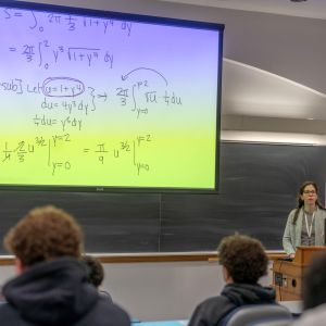 Professor teaching calculus in a lecture hall with equations on a large screen.