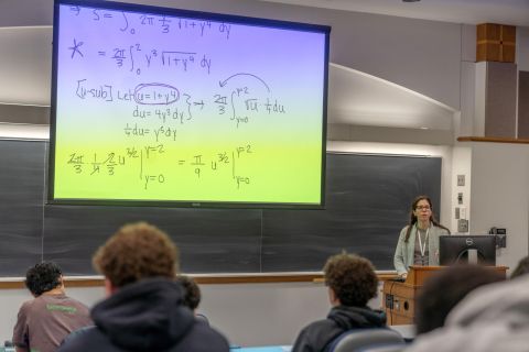 Professor teaching calculus in a lecture hall with equations on a large screen.