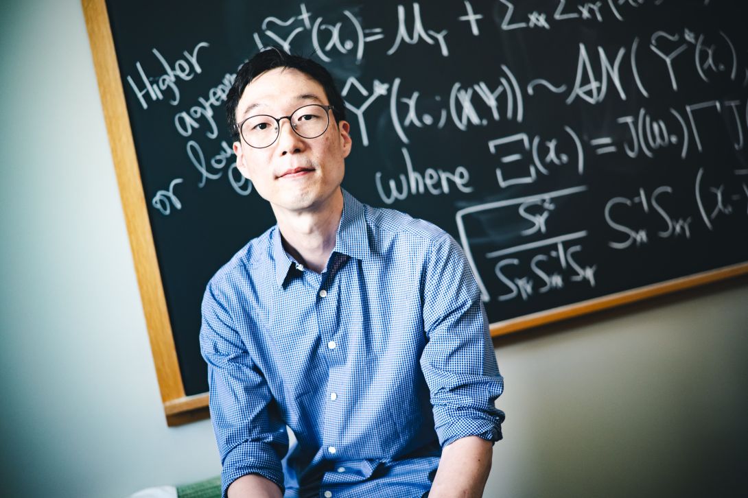 Taeho Kim wearing a blue button-up shirt and glasses sitting in front of a blackboard with math problems on it.