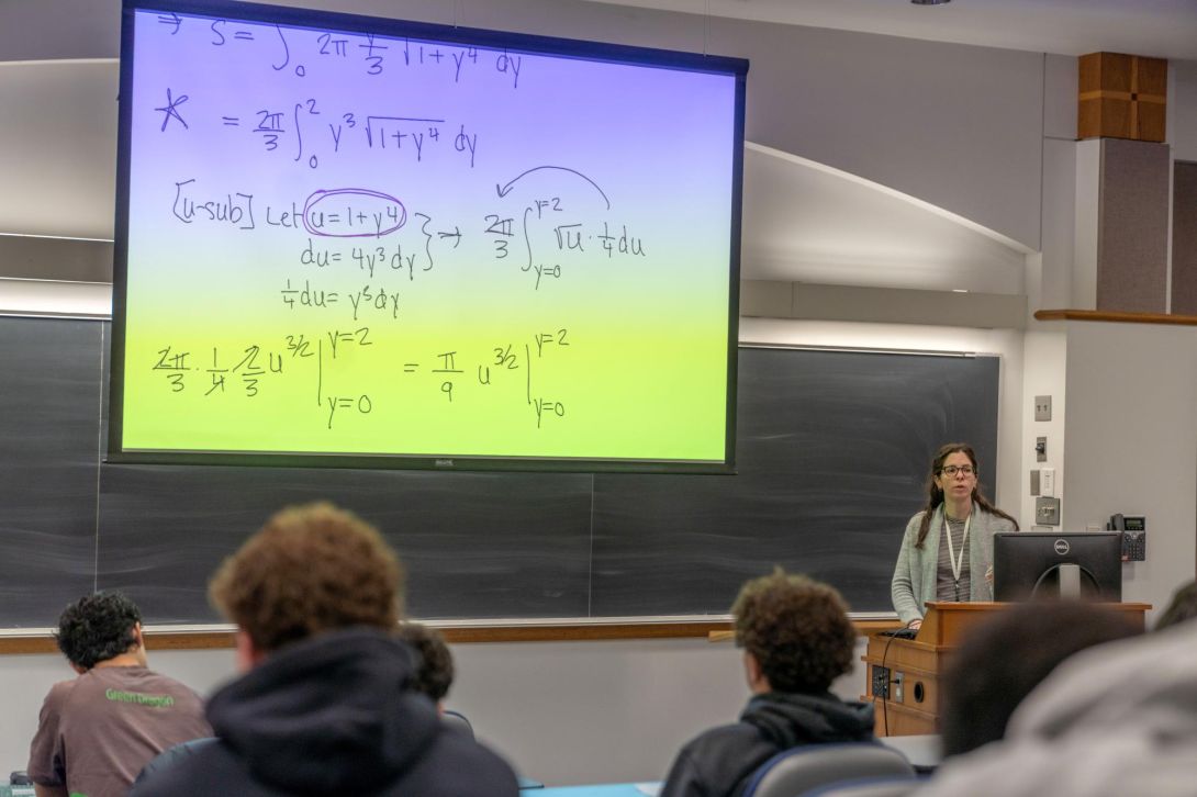 Professor teaching calculus in a lecture hall with equations on a large screen.